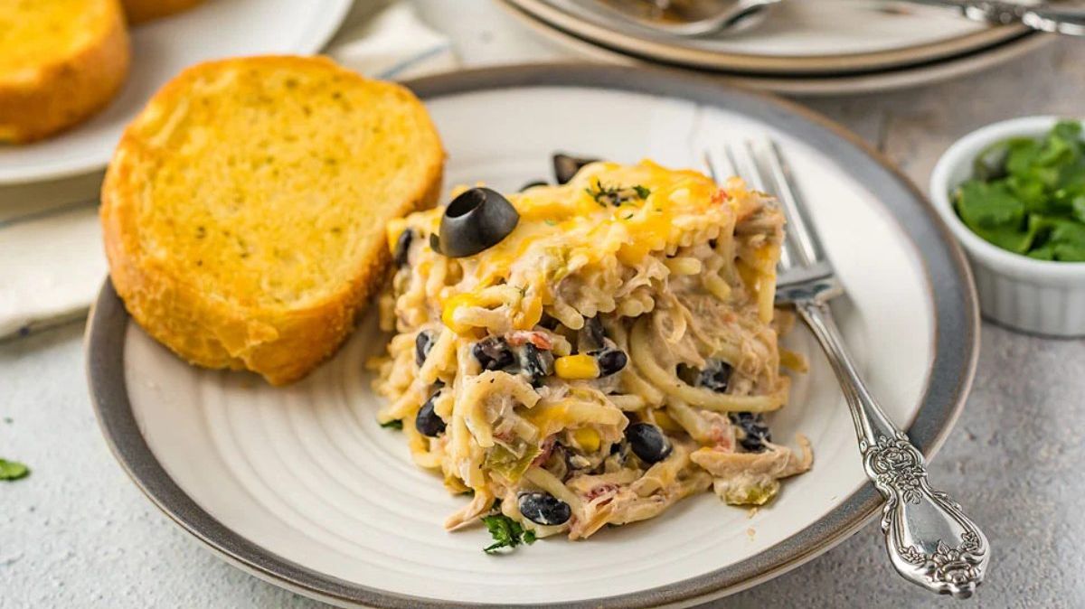 A serving of creamy chicken spaghetti with black olives and cheese on a plate, accompanied by a slice of toasted bread and a fork.