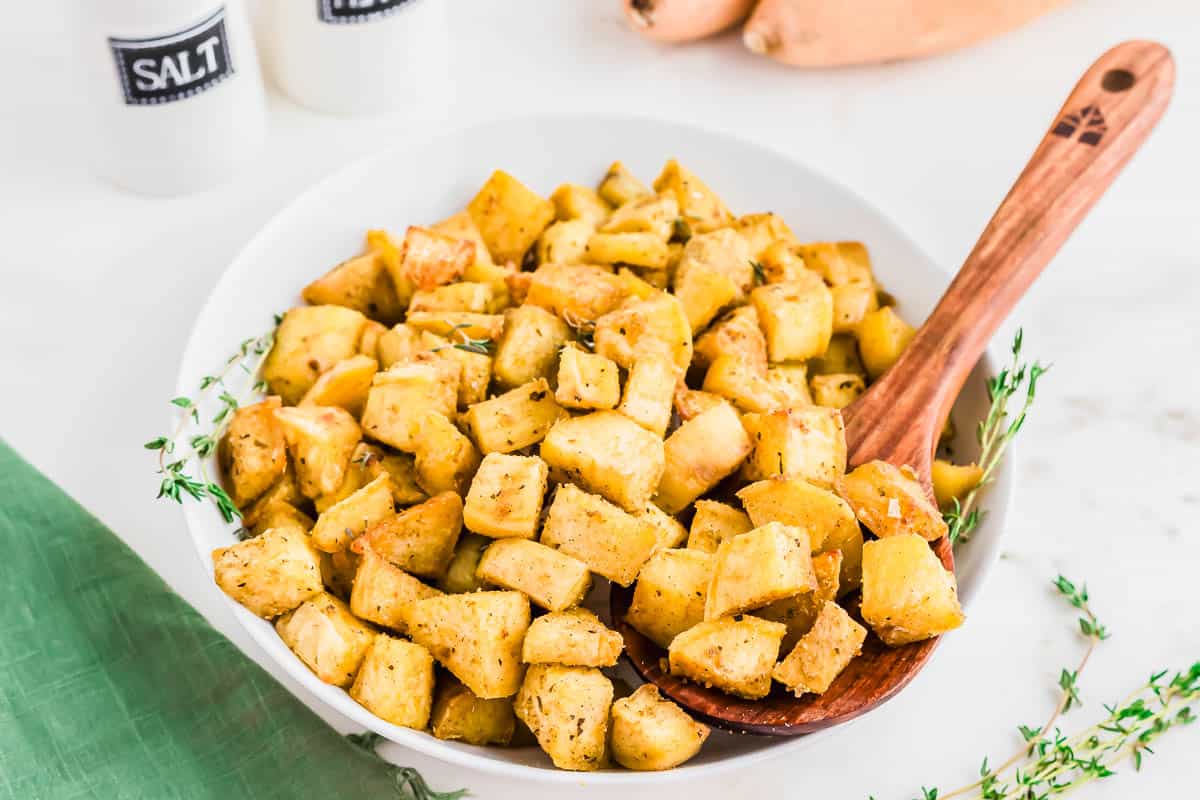 A white bowl filled with seasoned roasted butternut squash cubes, garnished with herbs, sits on a white surface with a wooden spoon.