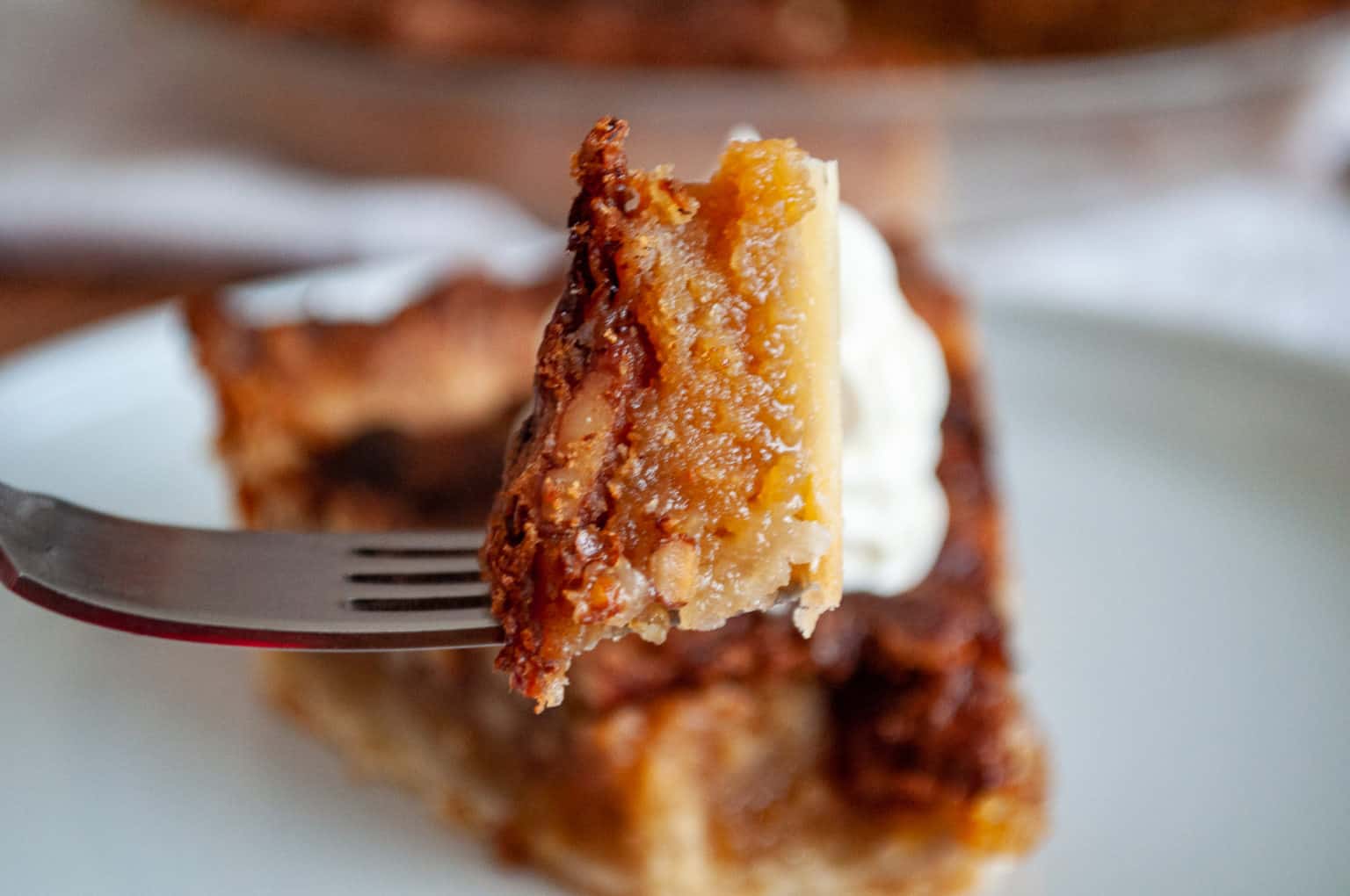 A close-up of a fork holding a bite of pie with a golden, gooey filling and flaky crust, with a dollop of whipped cream visible in the background.