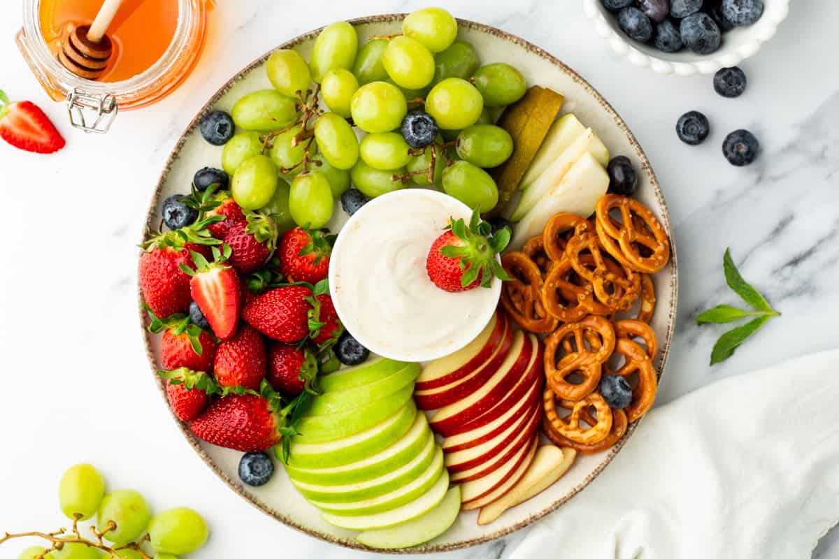 A round platter with green grapes, red and green apple slices, strawberries, blueberries, pretzels, pear slices, and a bowl of dip in the center.