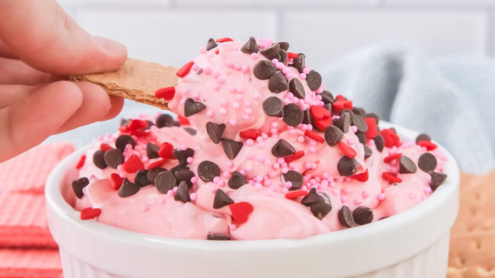 A hand dips a graham cracker into a bowl of pink whipped dessert topped with chocolate chips and red and pink sprinkles.