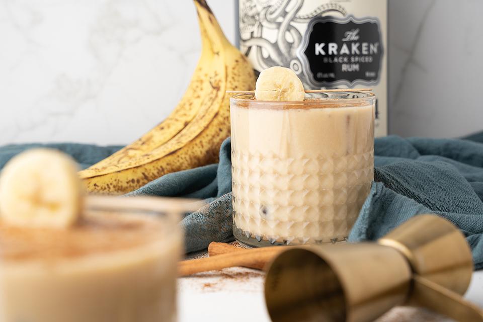 A glass of creamy banana cocktail garnished with a banana slice, with a ripe banana, bottle of Kraken Black Spiced Rum, and a jigger in the background.