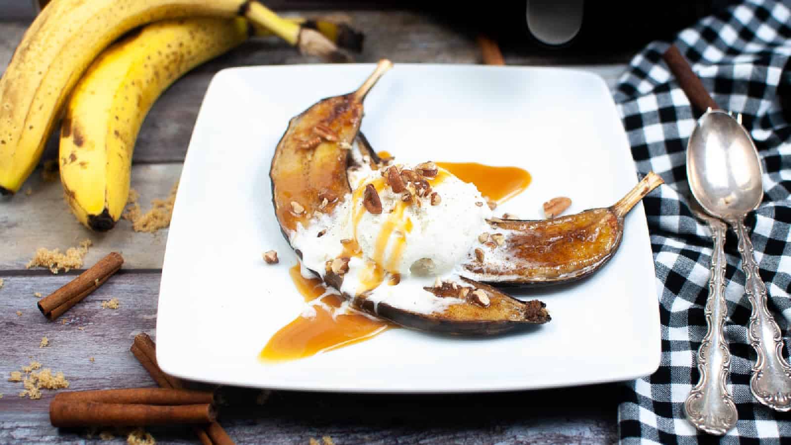A plate with baked banana halves topped with vanilla ice cream, caramel sauce, and chopped nuts, beside whole bananas, cinnamon sticks, and a checkered napkin with two spoons.