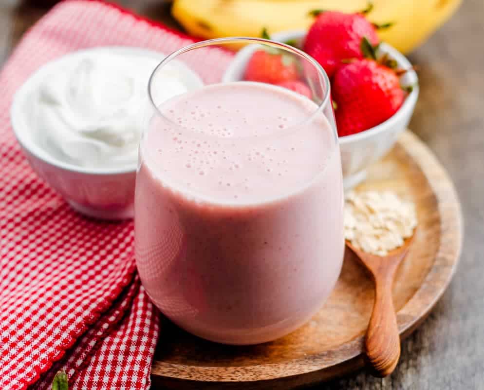 A glass of pink smoothie on a wooden plate, with a bowl of yogurt, a bowl of strawberries, a spoon of oats, and a red checkered cloth in the background.
