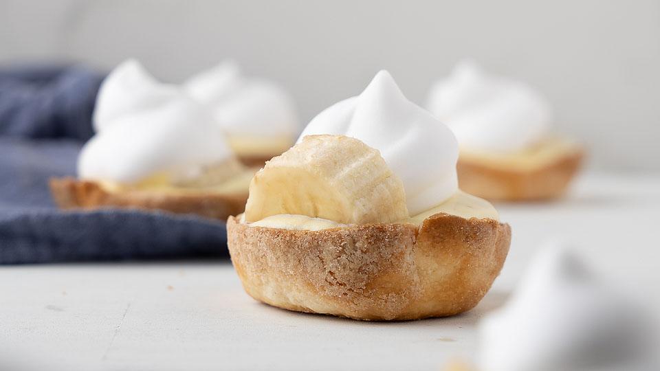 A small tart topped with a banana slice and a dollop of whipped cream sits on a white surface, with more tarts blurred in the background.