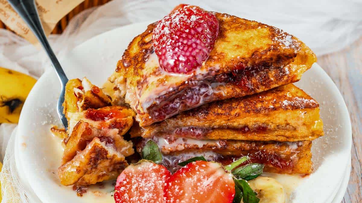 Stacked French toast sandwiches filled with cream and strawberries, dusted with powdered sugar, on a white plate with fresh strawberries and a forkful taken.