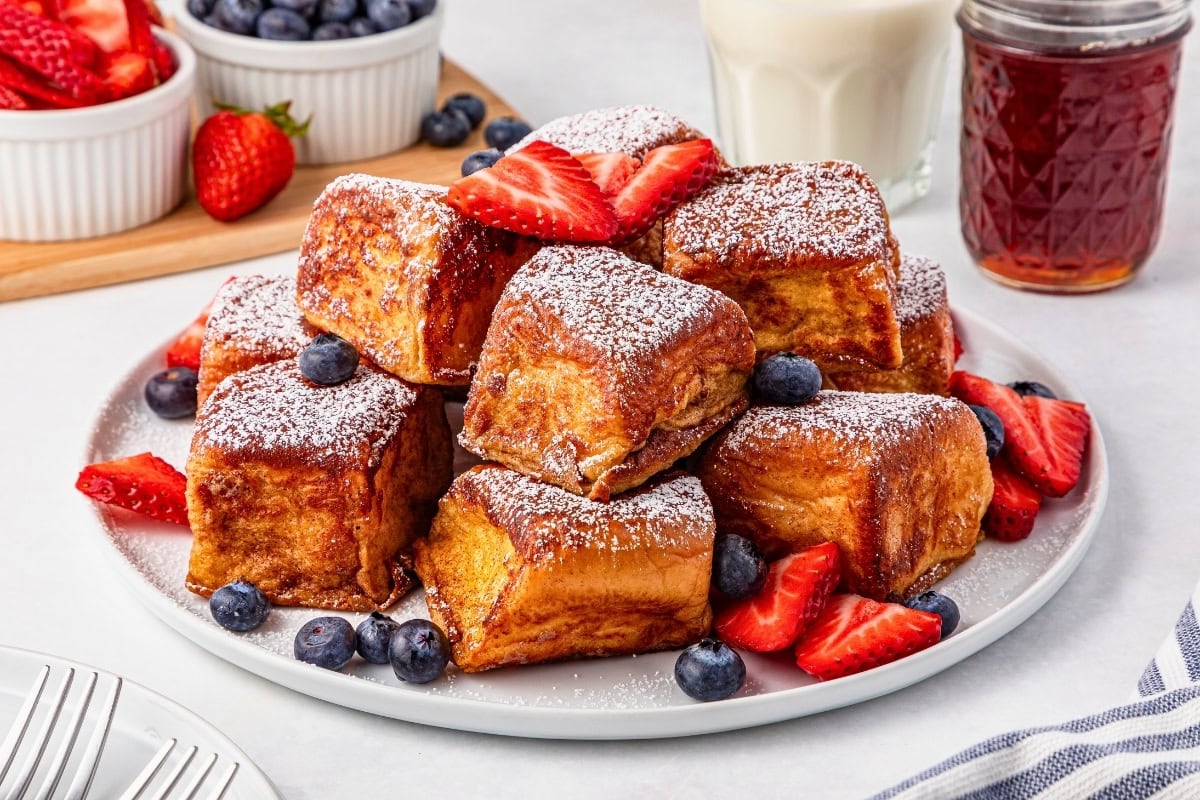 A plate of thick French toast cubes dusted with powdered sugar, garnished with fresh strawberries and blueberries, with a jar of jam and a glass of milk in the background.