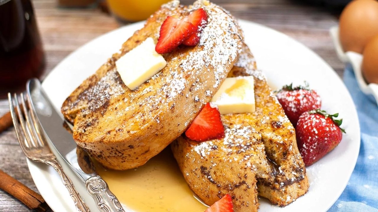 Two thick slices of French toast topped with butter, powdered sugar, and sliced strawberries on a white plate, with syrup and whole strawberries on the side.