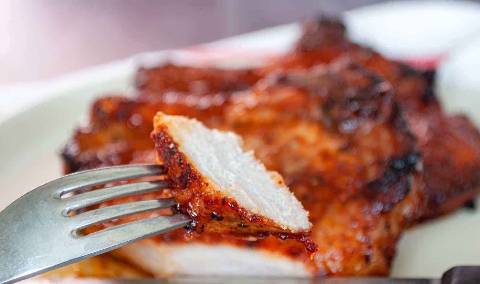 A close-up of a fork holding a piece of grilled, seasoned chicken breast, with more chicken pieces blurred in the background on a plate.
