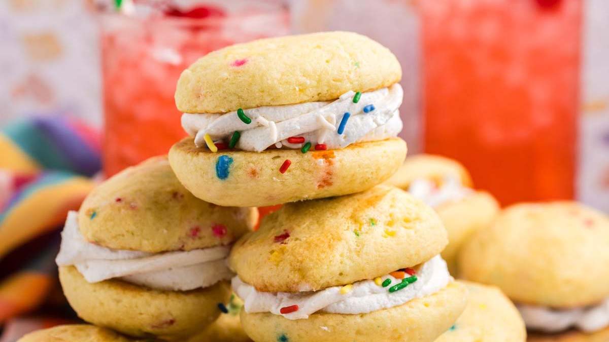 A stack of funfetti sandwich cookies with white frosting and colorful sprinkles, with more cookies and two pink drinks in the background.
