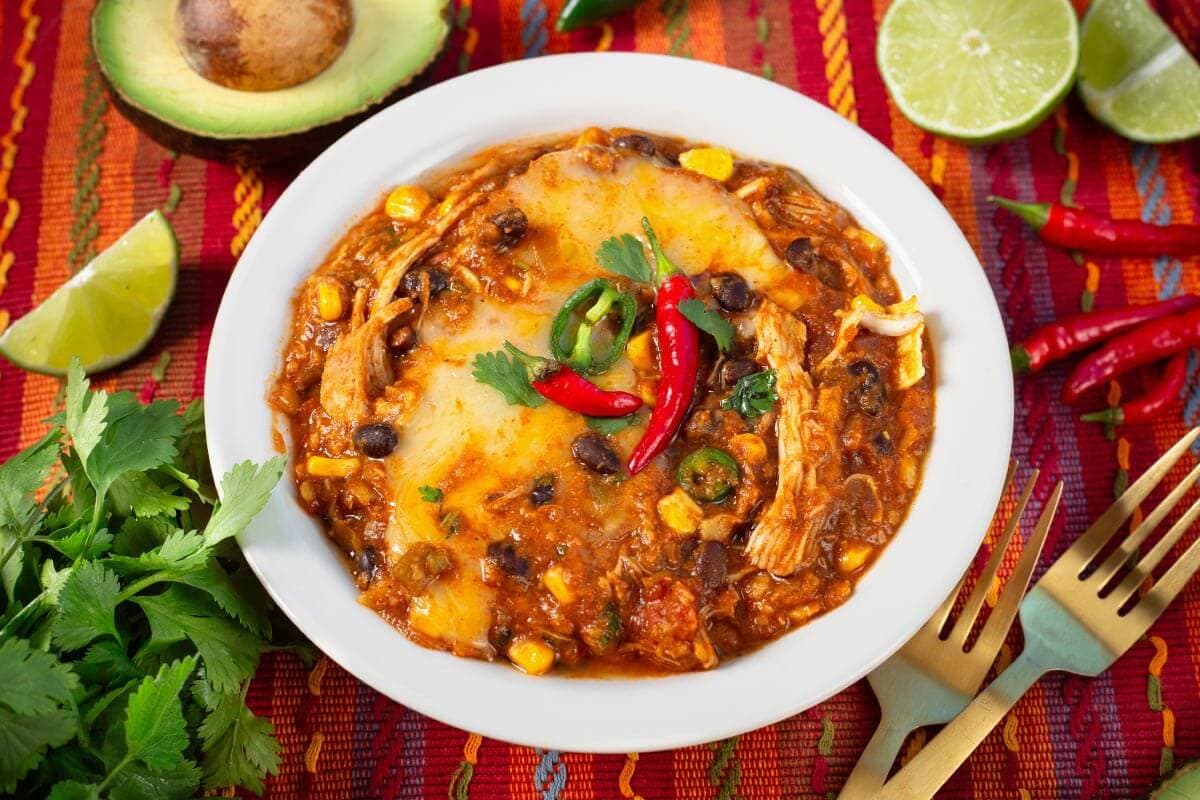 A bowl of chicken enchilada soup topped with cheese, cilantro, and red chili peppers, surrounded by avocado, lime wedges, cilantro, and red chilies on a colorful tablecloth.