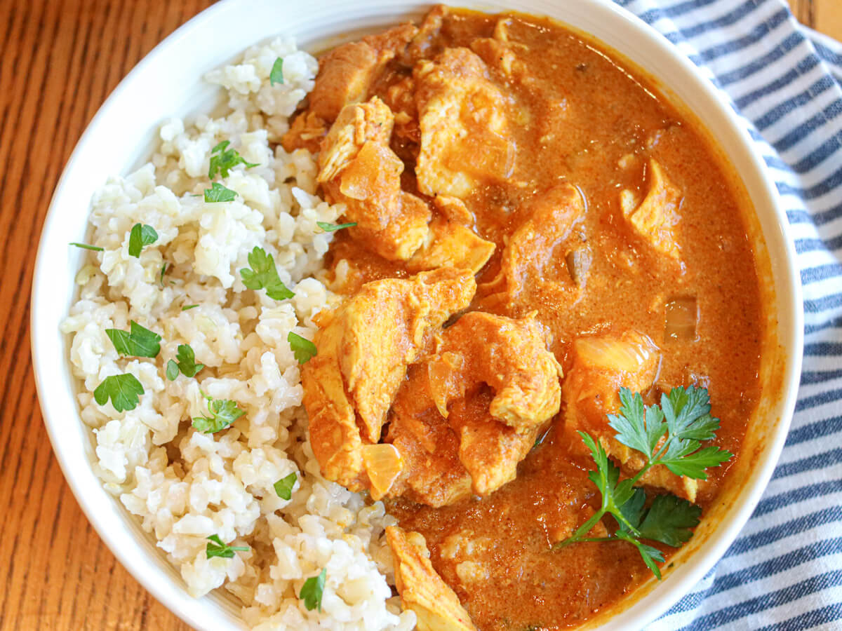 A bowl of brown rice and chicken curry garnished with fresh parsley, placed on a wooden table next to a striped cloth.