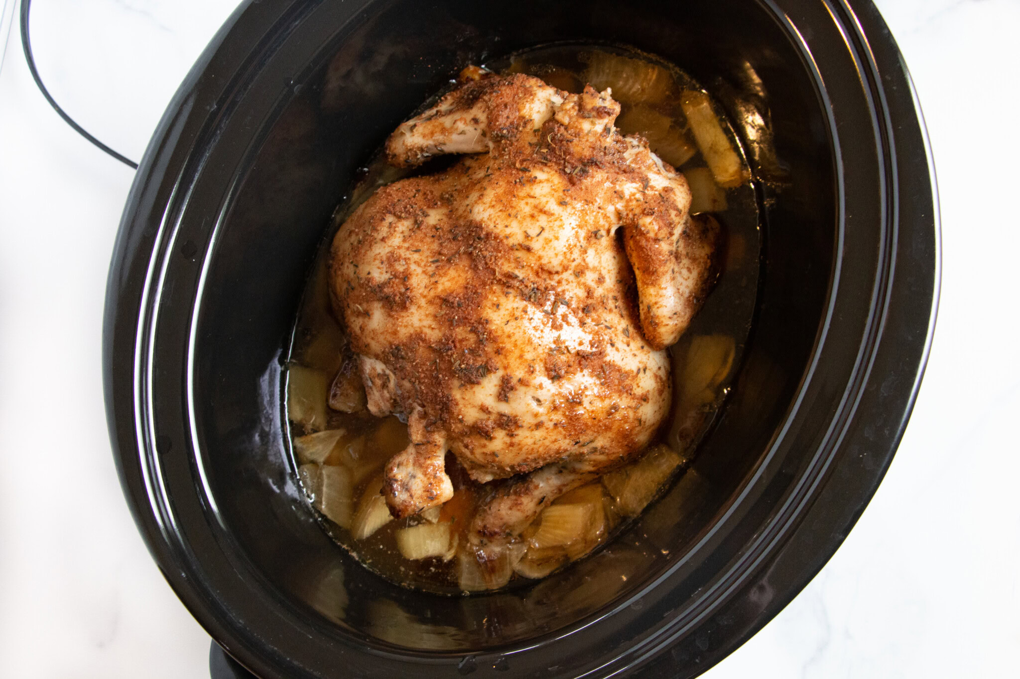 Whole seasoned chicken cooking in a black slow cooker with chopped onions and juices visible at the bottom.