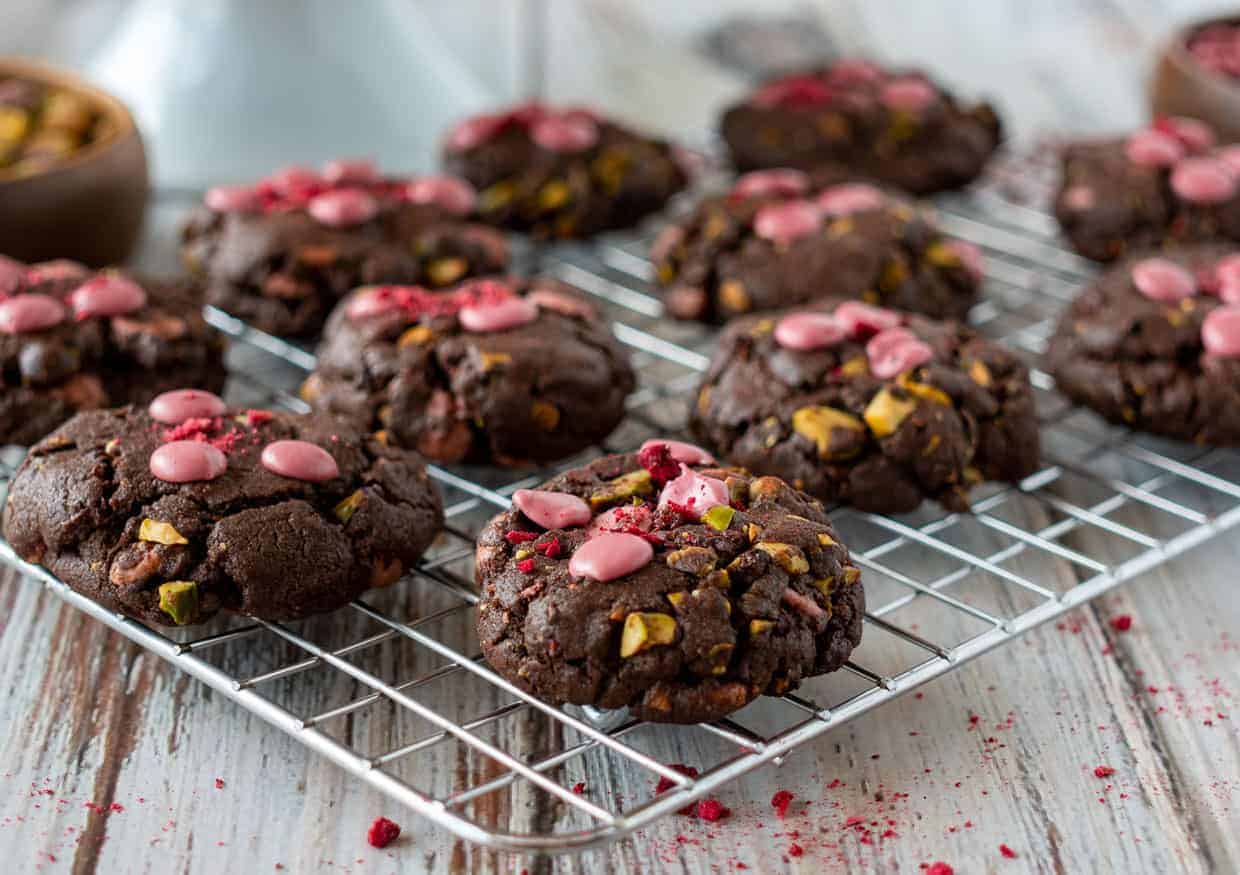 Chocolate cookies with pink chocolate chips and pistachio pieces are cooling on a metal wire rack, with crumbs scattered on a rustic wooden surface.
