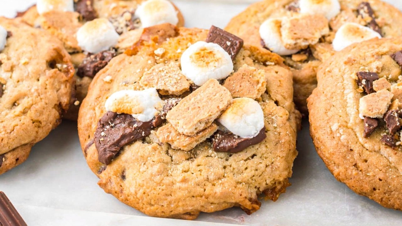Close-up of large cookies topped with toasted marshmallows, chocolate pieces, and crumbled graham crackers on a white surface.