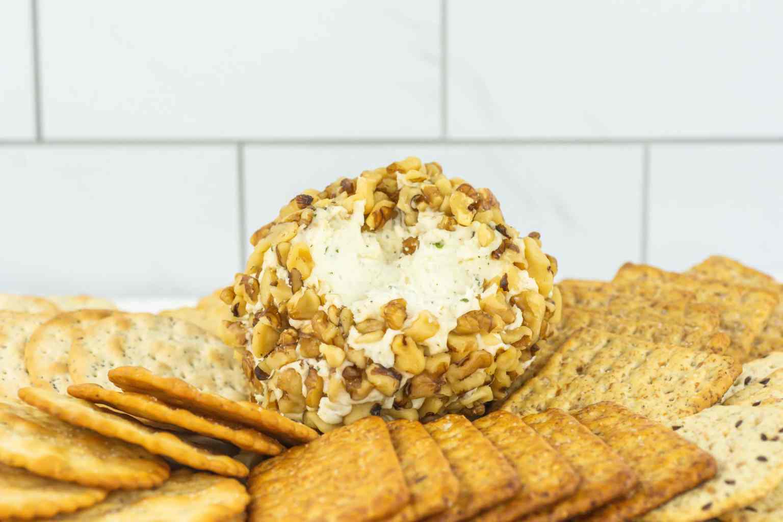 A cheese ball coated in chopped nuts is surrounded by assorted crackers on a white surface with a tiled background.