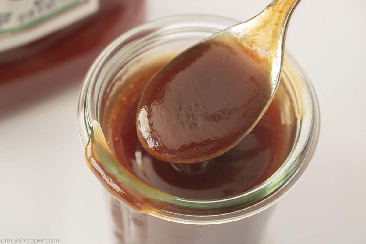 A spoon with thick barbecue sauce hovers over an open glass jar filled with barbecue sauce.