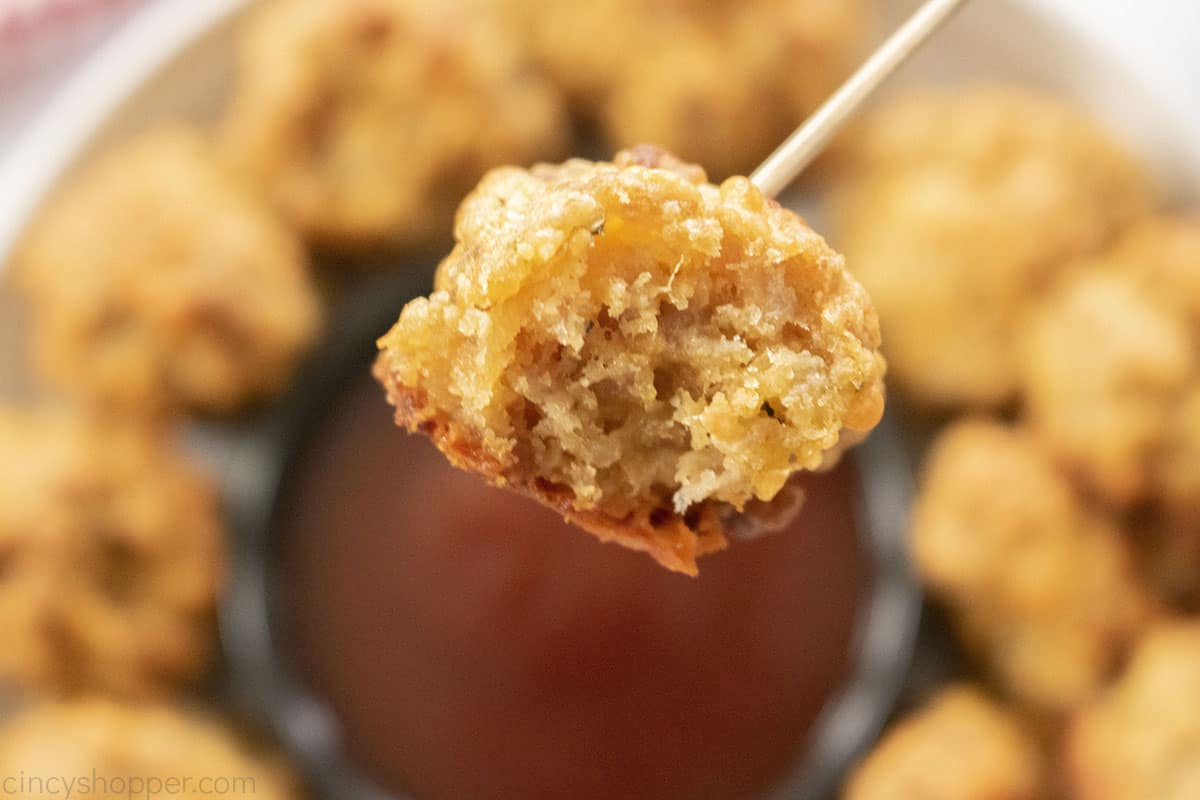 A close-up of a breaded, bite-sized appetizer on a toothpick with a bowl of red dipping sauce and more appetizers in the background.