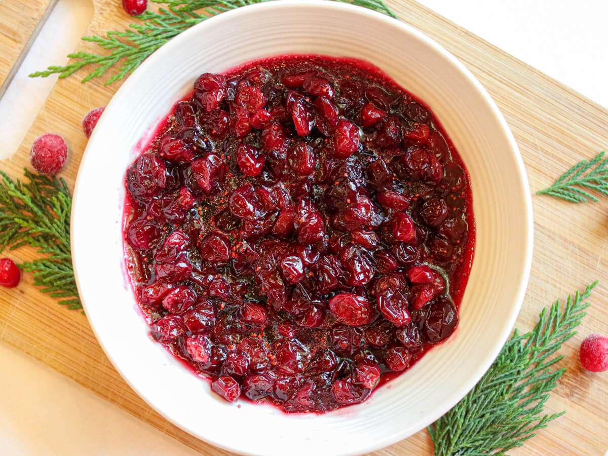 A bowl of cranberry sauce placed on a wooden board, garnished with cranberries and evergreen sprigs.