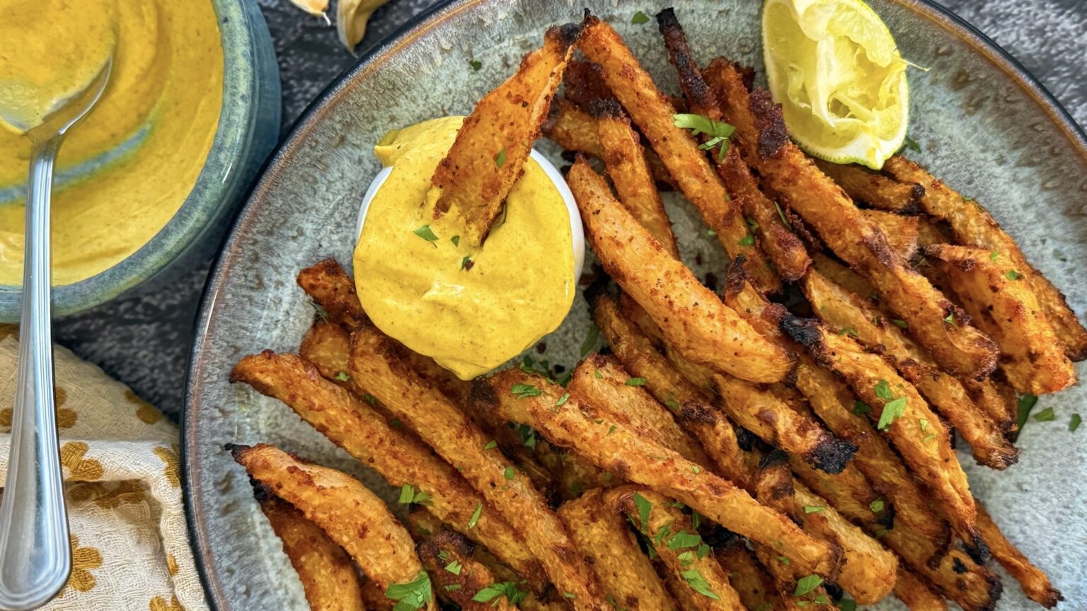 A plate of seasoned, crispy fries is served with a yellow dipping sauce and a wedge of lemon.