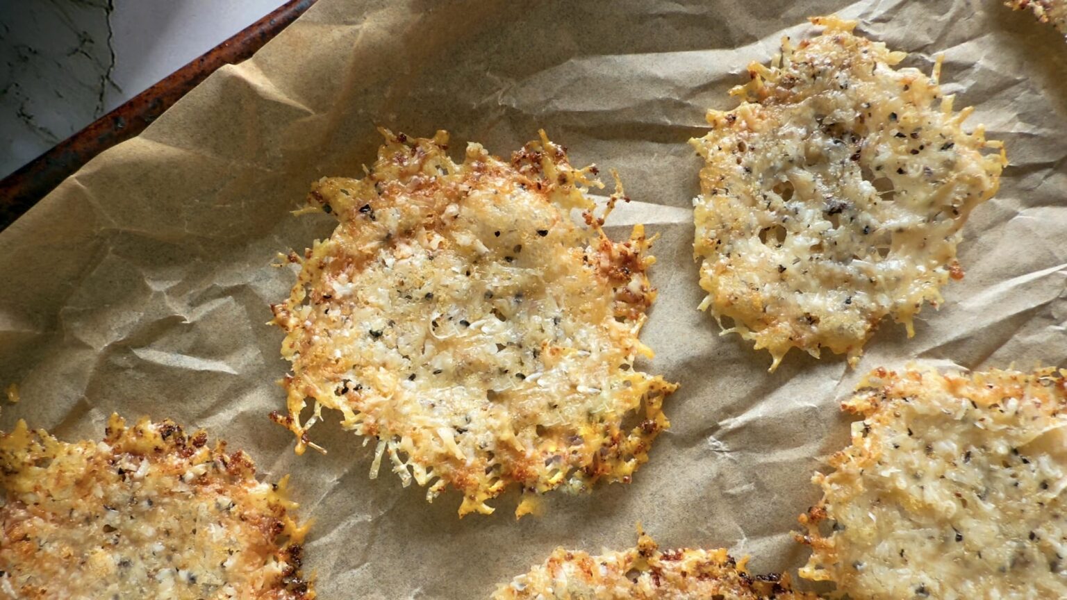 Baked cheese crisps with seasoning on parchment paper, shown on a baking tray.