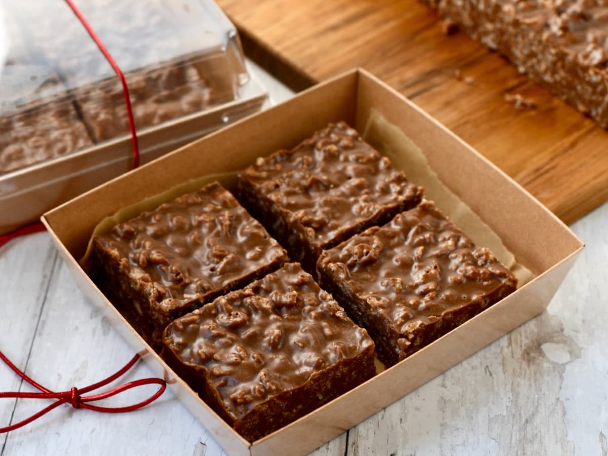 A cardboard box containing four chocolate rice crispy treats sits on a white surface, with more treats and a closed box in the background.