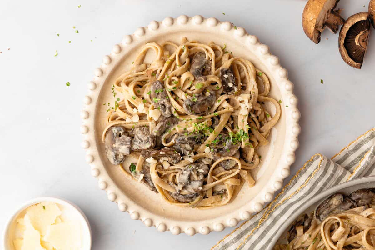 A plate of fettuccine pasta with creamy mushroom sauce, garnished with chopped parsley and grated cheese, on a white table.