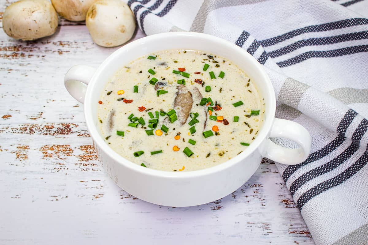 A white bowl of creamy mushroom soup garnished with chopped chives and red pepper flakes, placed on a rustic white table with a striped towel and whole mushrooms nearby.