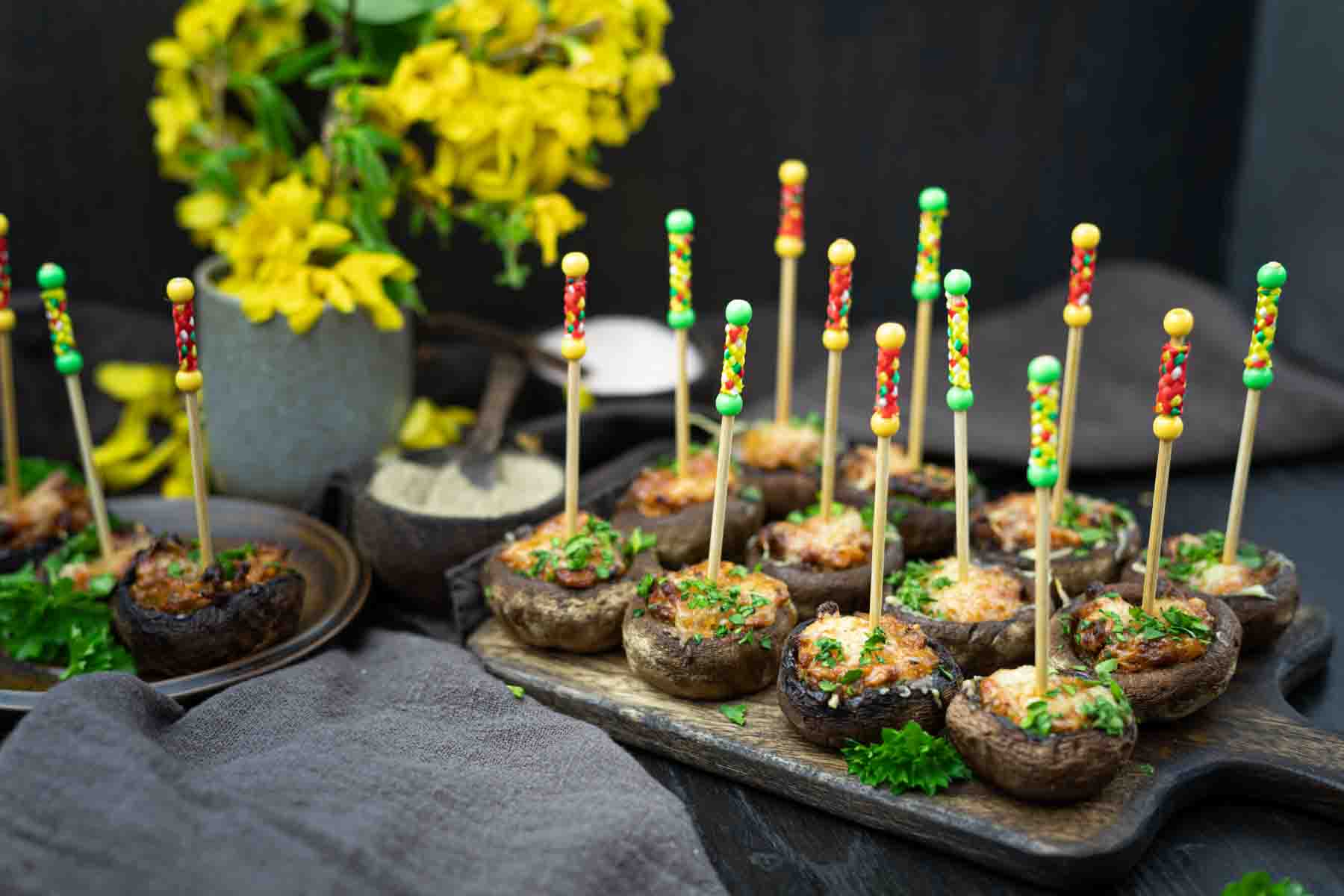 Stuffed mushrooms on platters, each with a colorful cocktail stick, garnished with parsley. A vase with yellow flowers is in the background.