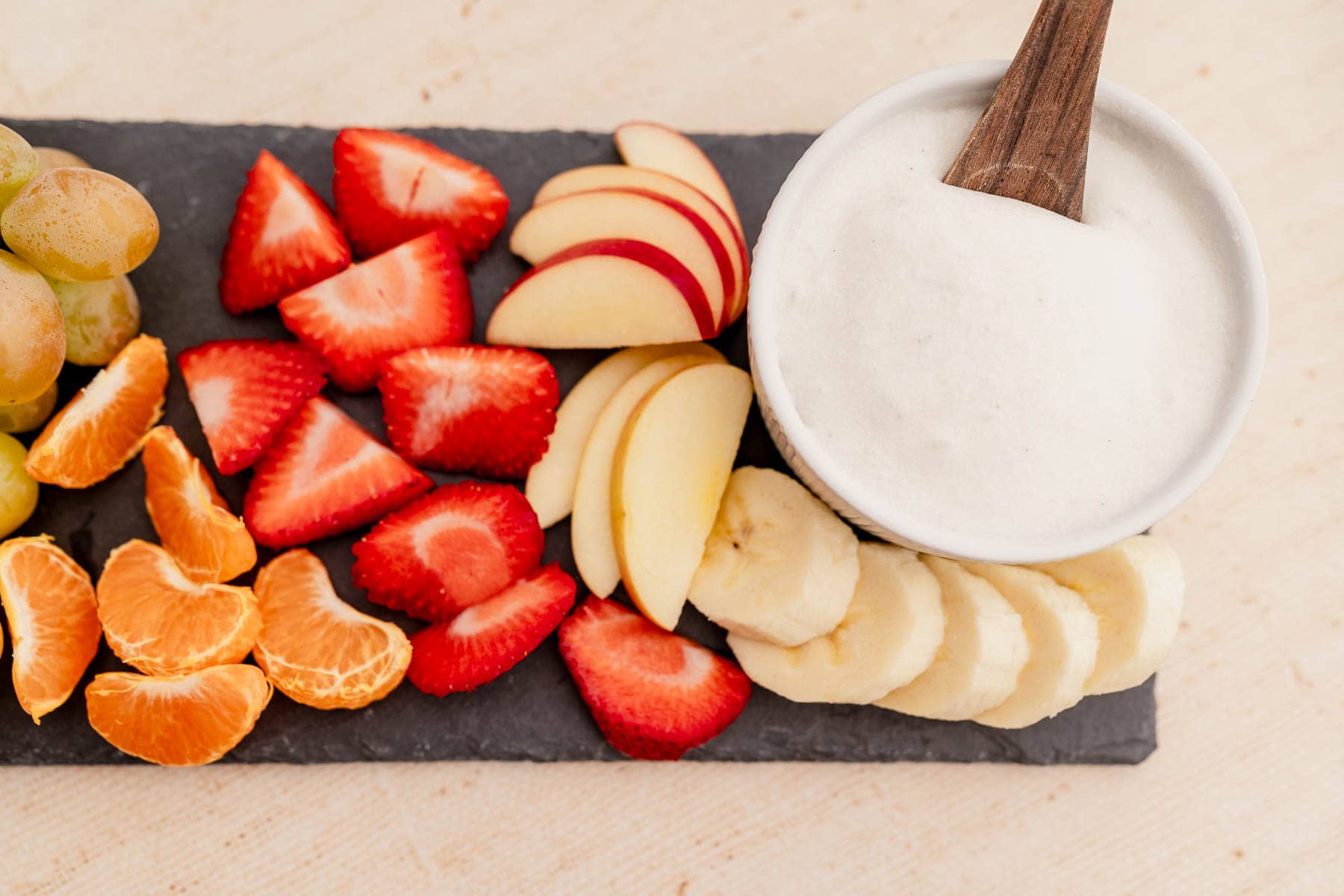 Sliced strawberries, apples, bananas, mandarin orange segments, grapes, and a bowl of white yogurt with a wooden spoon on a slate tray.