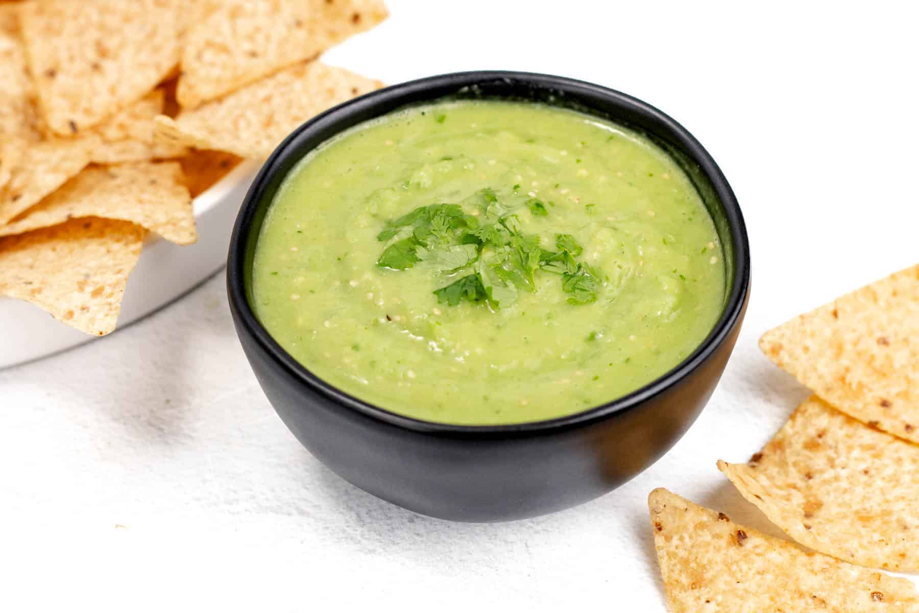 A black bowl of green guacamole garnished with cilantro is surrounded by tortilla chips on a white surface.