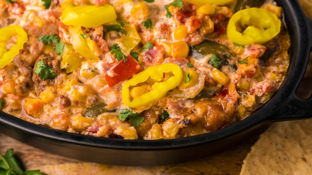 A close-up of a cheesy dip with corn, diced tomatoes, green peppers, and sliced yellow banana peppers in a black dish.