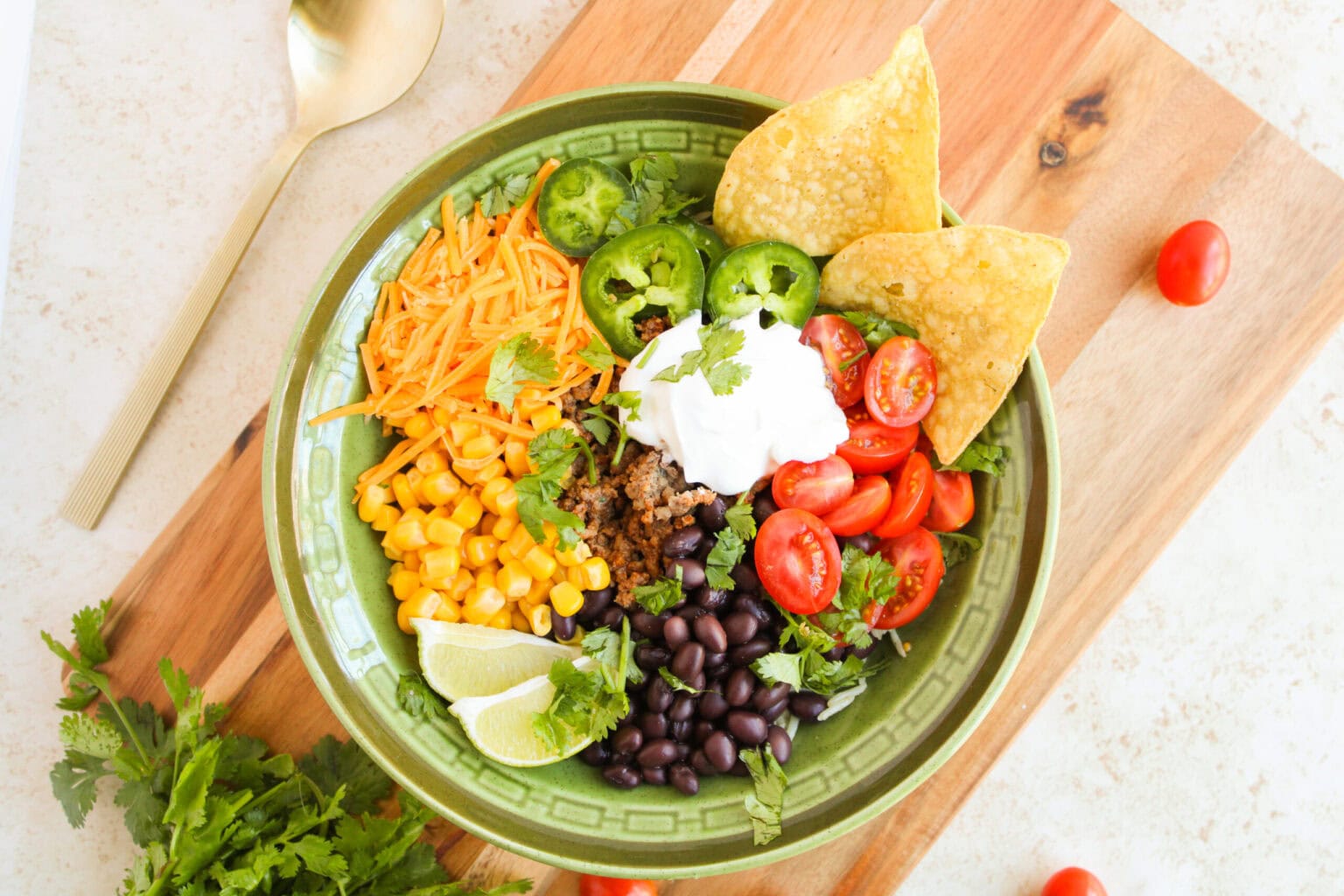 A green bowl filled with ground meat, black beans, corn, shredded cheese, cherry tomatoes, jalapeรฑo slices, tortilla chips, lime wedges, sour cream, and cilantro on a wooden board.