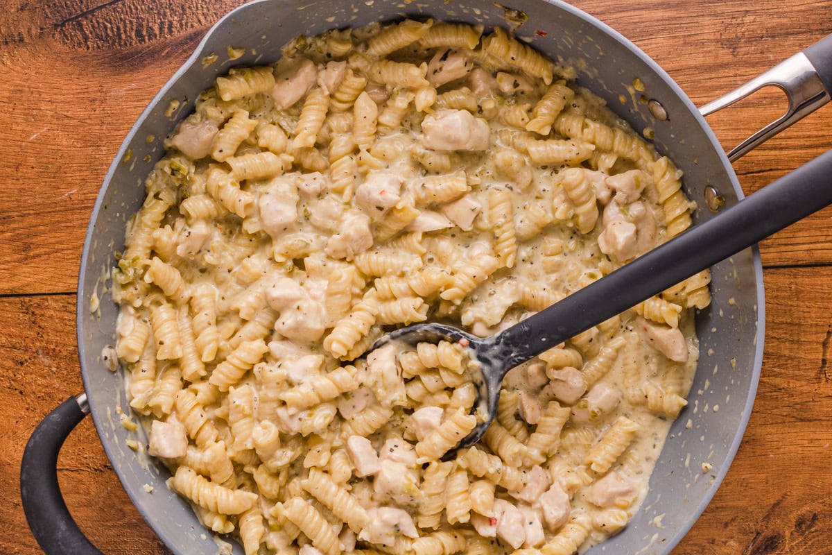 A skillet filled with creamy pasta and chunks of chicken, mixed with a creamy sauce and herbs, with a black serving spoon inside.