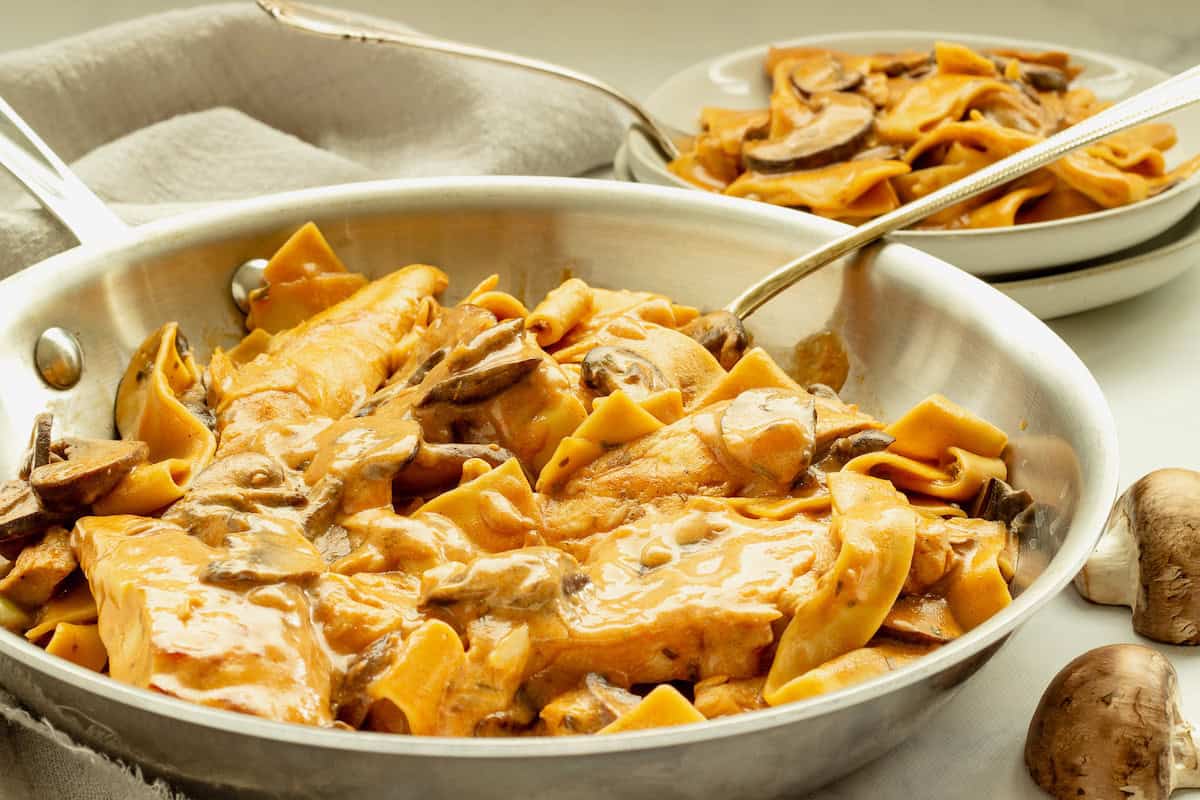 Wide pasta noodles with mushrooms in a creamy sauce served in a metal pan, with an additional portion on stacked plates in the background.