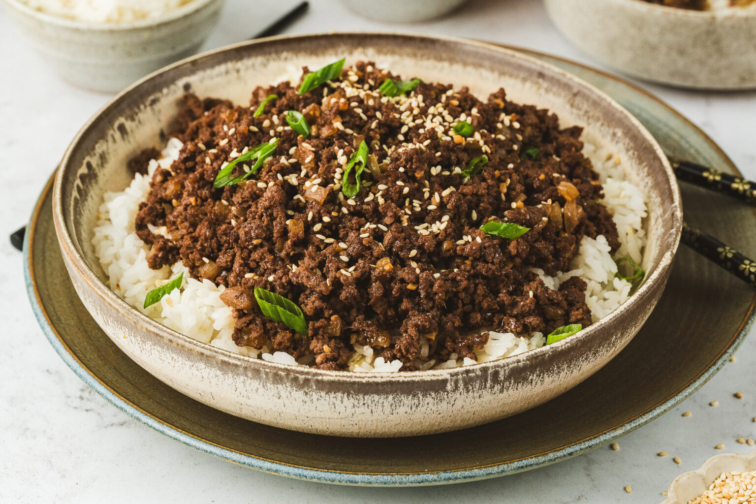 A bowl of white rice topped with seasoned ground beef, garnished with sesame seeds and chopped green onions.
