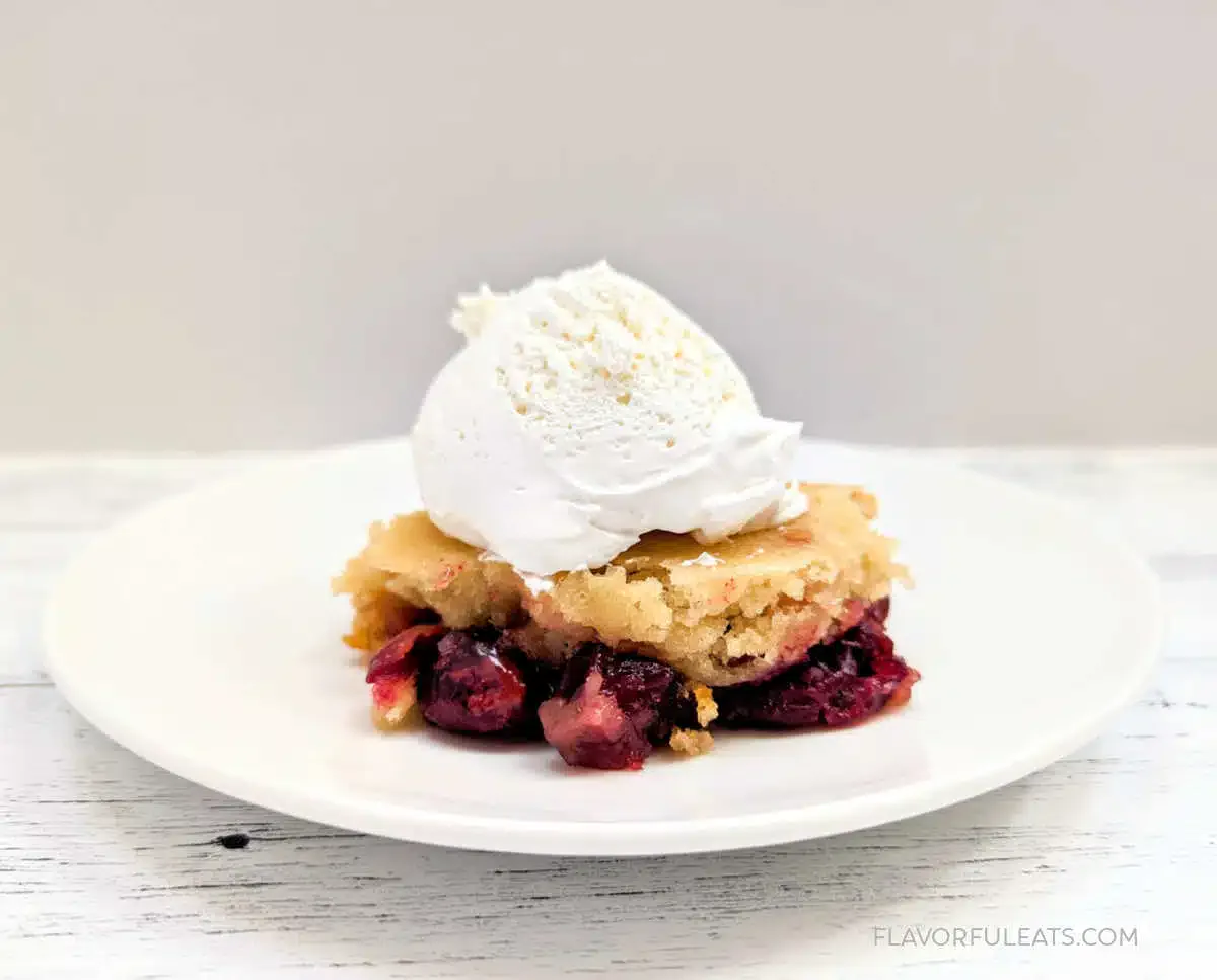 A serving of cherry dump cake topped with a scoop of vanilla ice cream is on a white plate against a plain background.