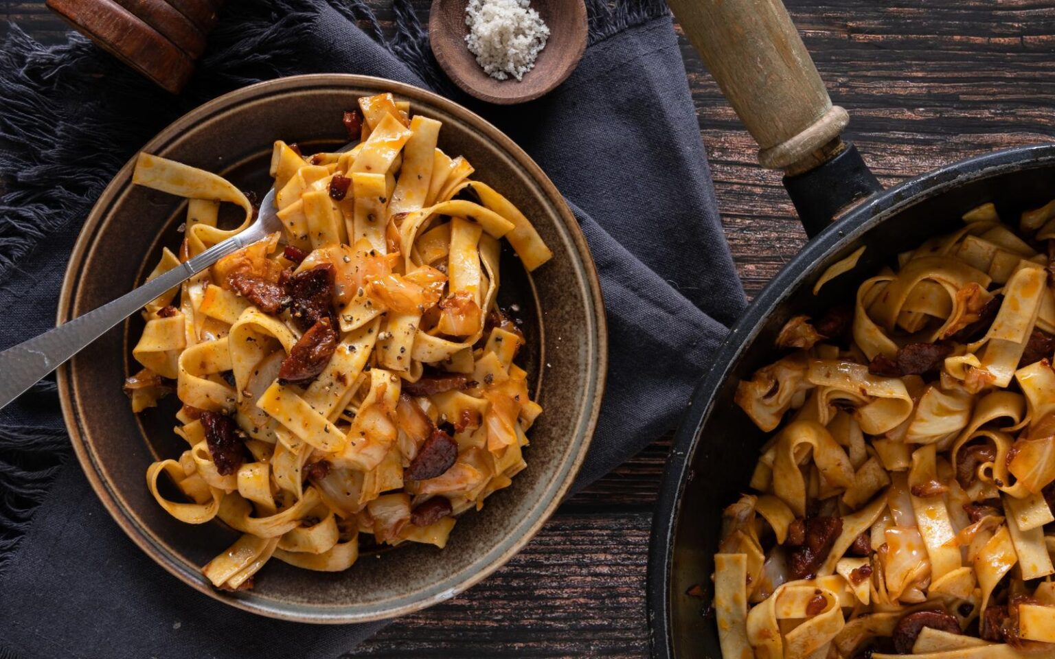 A bowl of tagliatelle pasta with tomato sauce and pieces of sausage, next to a pan of pasta and a small dish of salt on a dark wooden table.