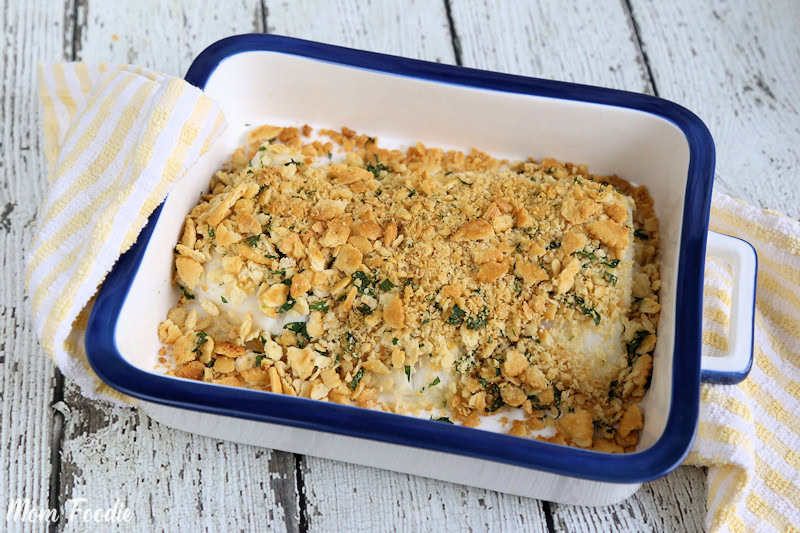 A baked fish fillet topped with a golden, crumbly cracker and herb crust in a white rectangular baking dish on a rustic wooden surface.