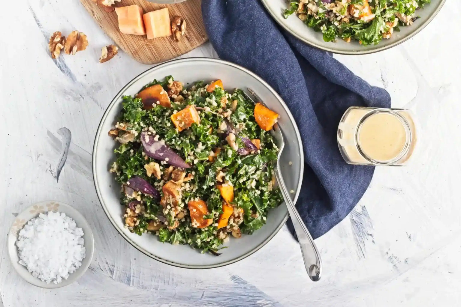 A bowl of kale salad with roasted vegetables, walnuts, and grains next to a fork, a small bowl of salt, dressing, and a cutting board with cheese cubes.