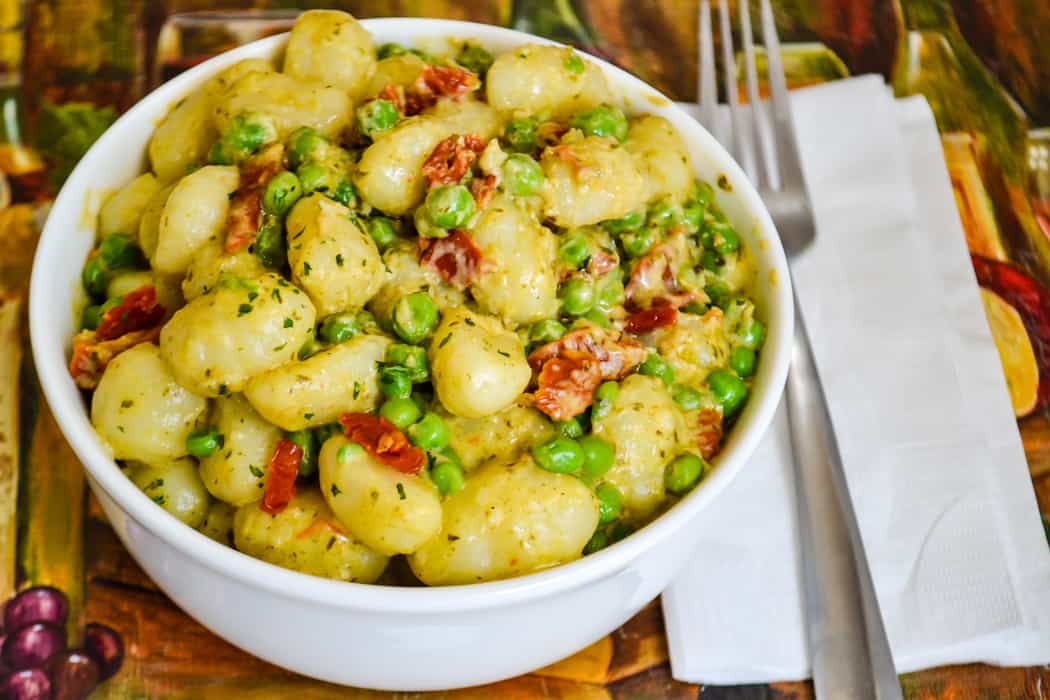 A white bowl filled with gnocchi, green peas, sun-dried tomatoes, and herbs sits on a table next to a fork, knife, and napkin.