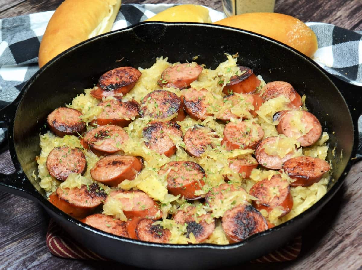 A cast iron skillet filled with sliced browned sausage mixed with sauerkraut, garnished with herbs; bread rolls and a checkered cloth are in the background.