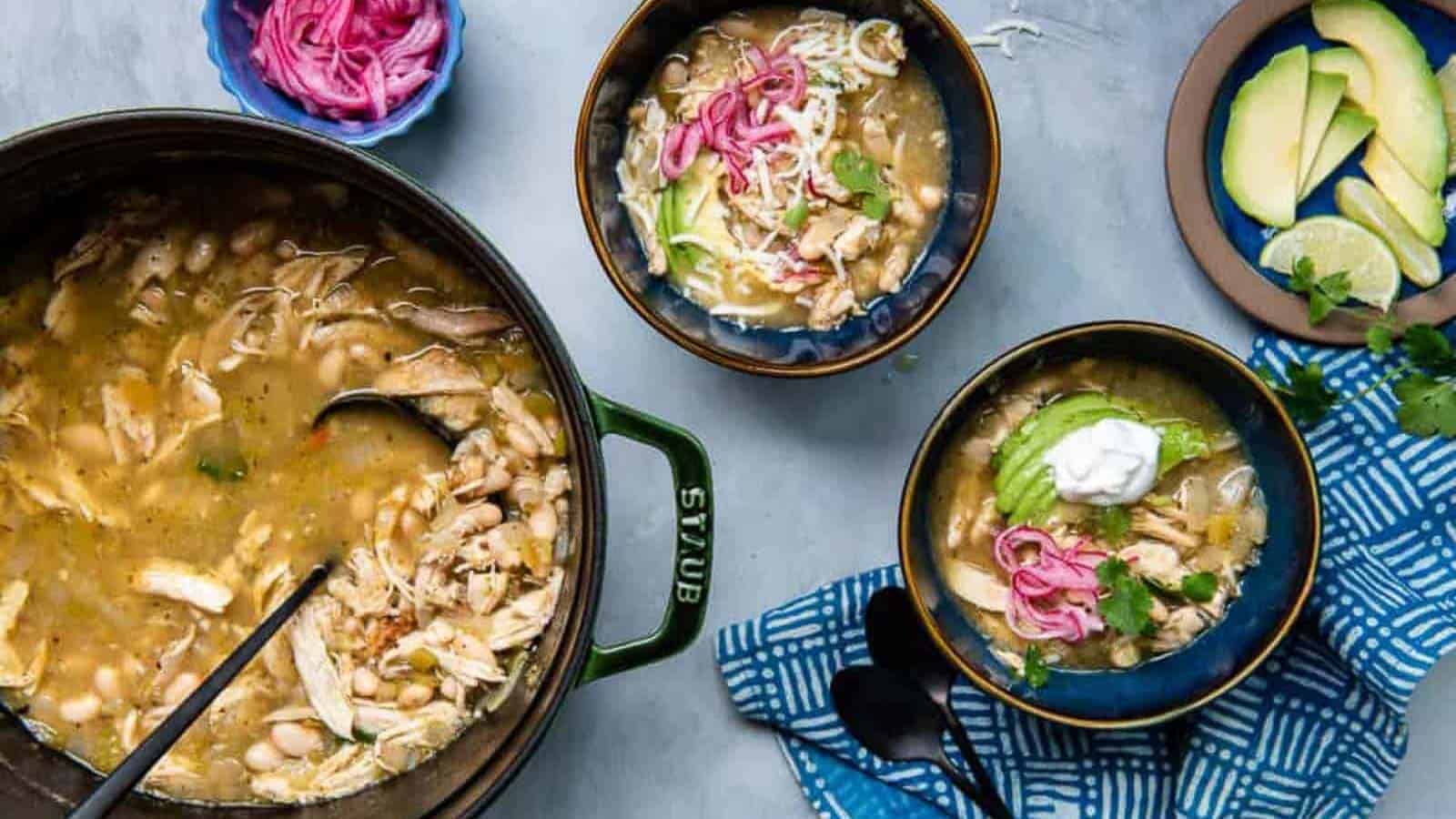 A pot of white chicken chili with two bowls served, topped with avocado, pickled onions, sour cream, and shredded cheese; sides include extra pickled onions and sliced avocado.