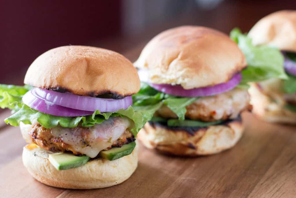 Three chicken burgers with lettuce, tomato, red onion, cheese, and avocado slices on toasted buns, arranged on a wooden surface.