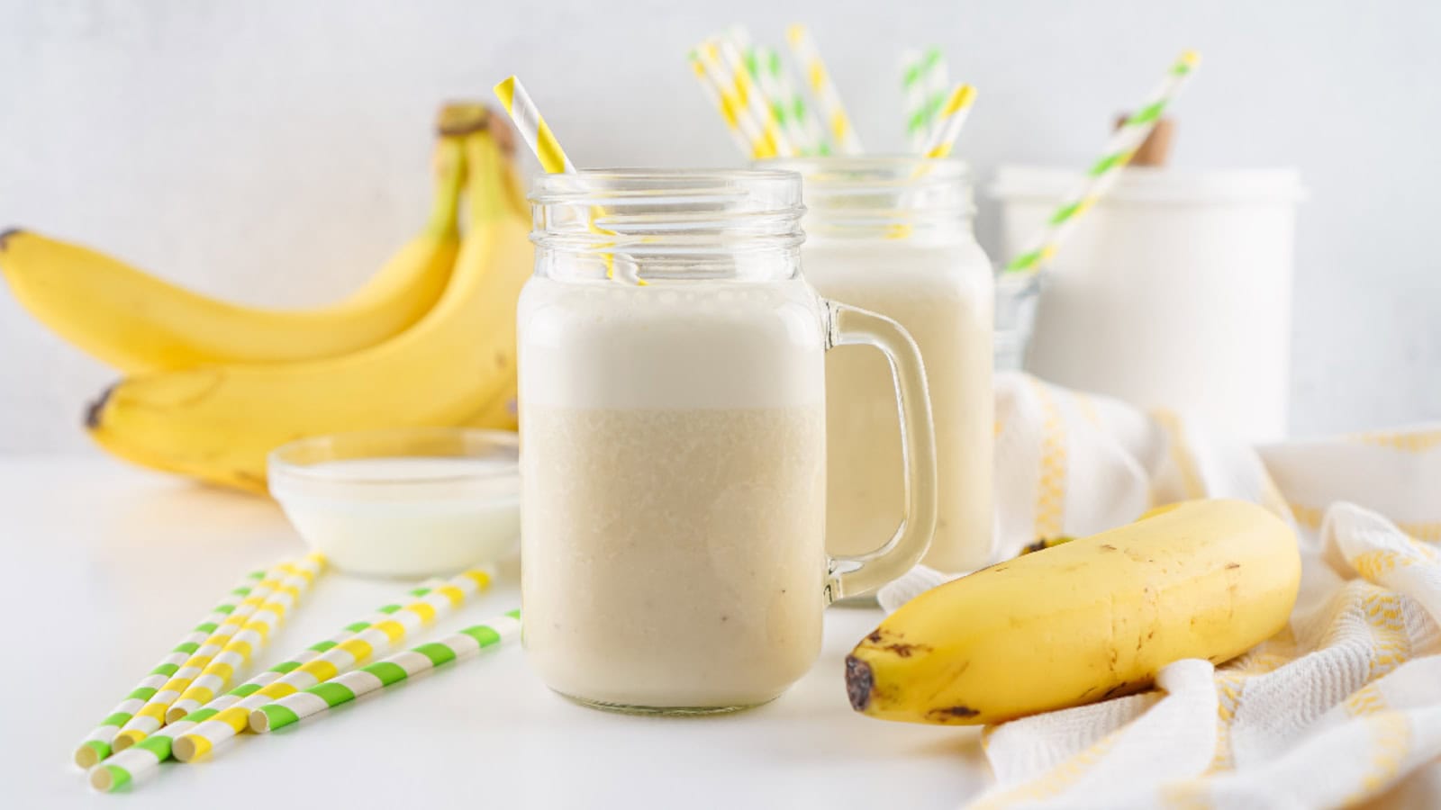 Two glass jars filled with banana smoothies, surrounded by fresh bananas, a bowl of yogurt, striped straws, and a white towel on a white surface.