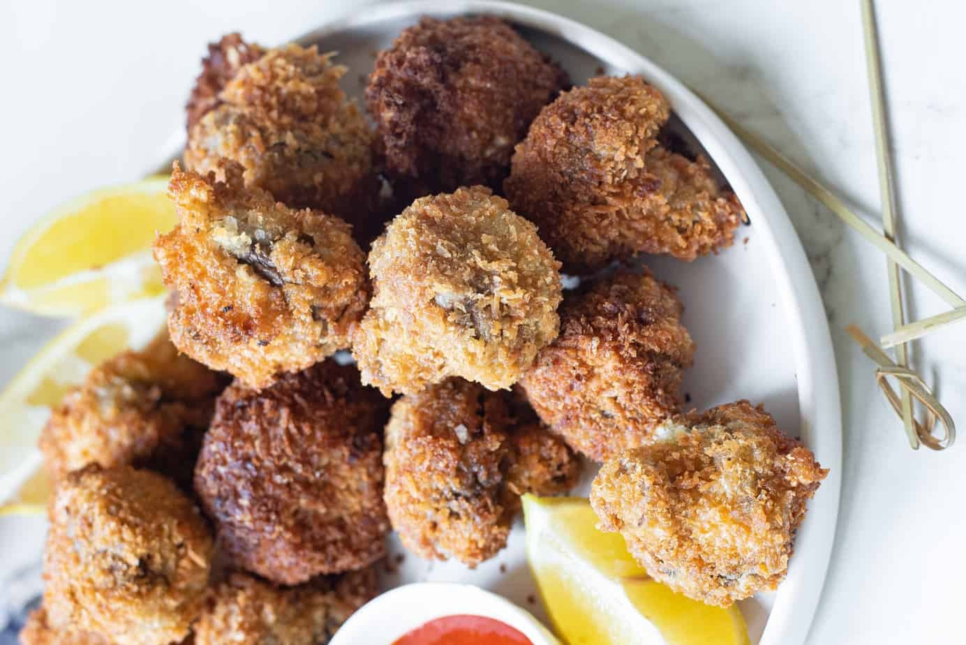 A plate of golden-brown fried mushrooms is served with lemon wedges and a small dish of red dipping sauce.