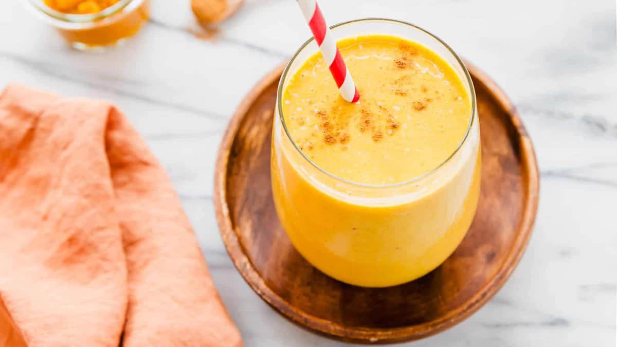 A glass of yellow smoothie with a red and white striped straw sits on a wooden plate, next to a peach-colored cloth on a marble surface.