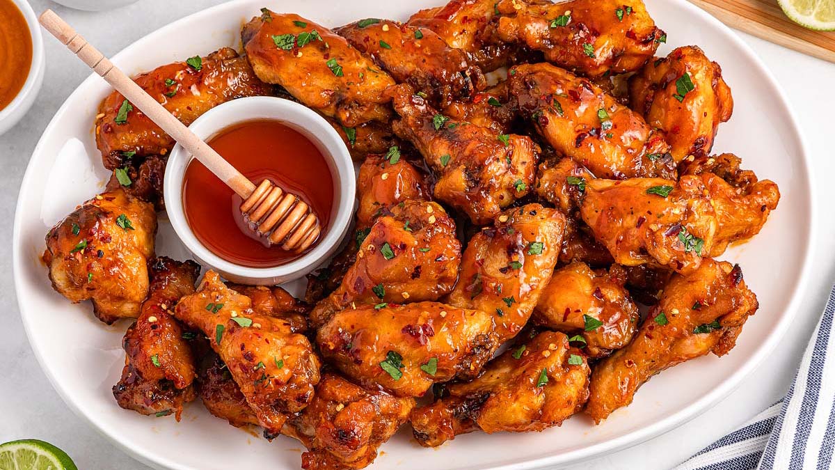 A platter of glazed chicken wings garnished with herbs, served with a small bowl of honey and a honey dipper.