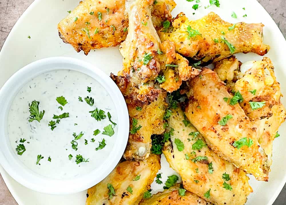 Plate of seasoned chicken wings garnished with chopped herbs, served with a side of creamy dipping sauce topped with parsley.