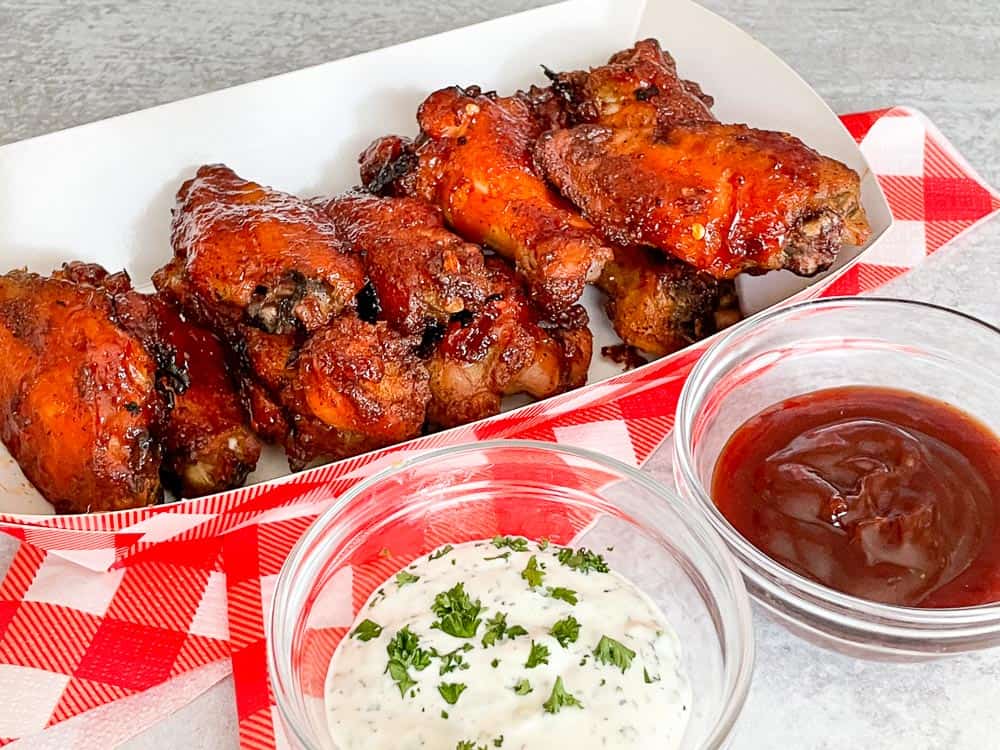 A tray of glazed chicken wings on a red and white checkered cloth with two small bowls of dipping sauces, one creamy with herbs and one dark red.